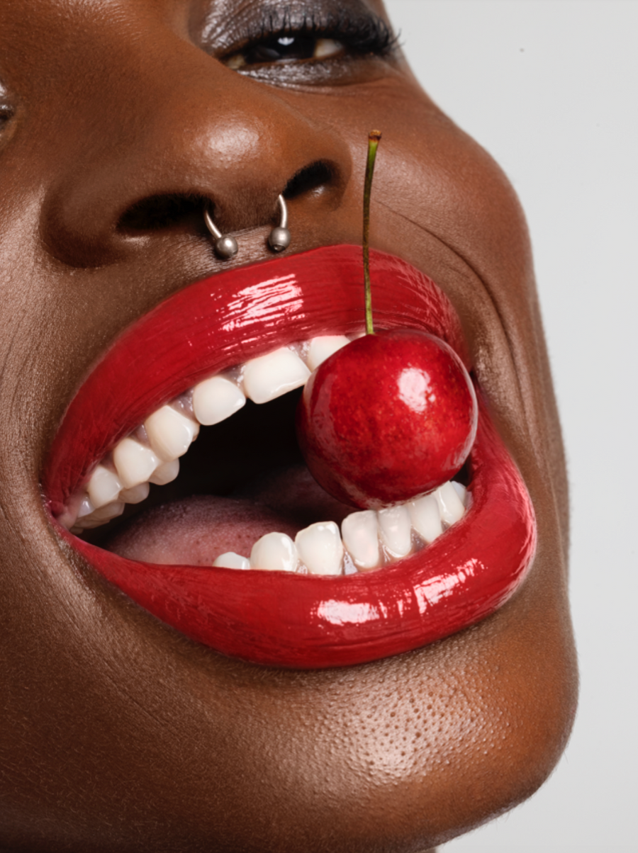 Close-up of a person with red lipstick holding a cherry in their mouth against a neutral background